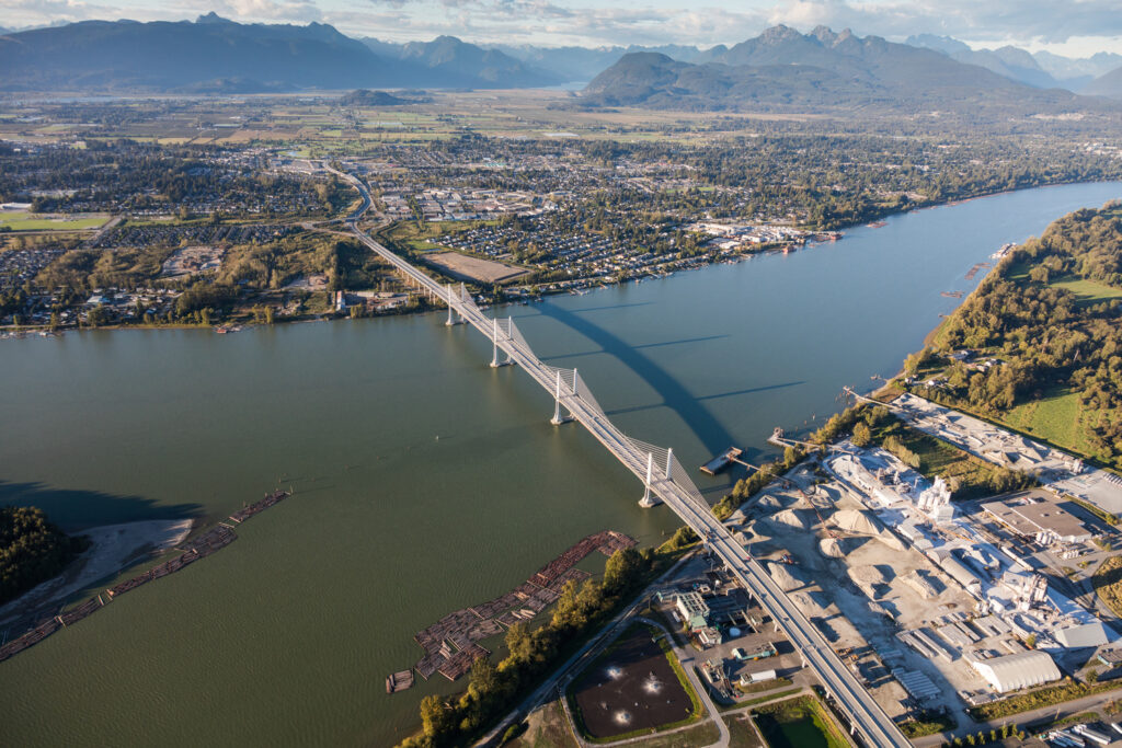 Aerial view of a river with a large cable-stayed bridge crossing it, surrounded by a landscape of mountains, forests, and an urban area.