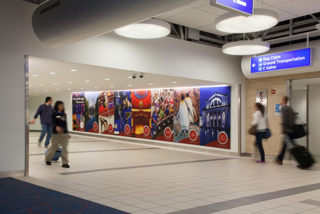 People walking past a series of colorful murals in an airport terminal. A sign overhead gives directions to baggage claim, ground transportation, and gates C.