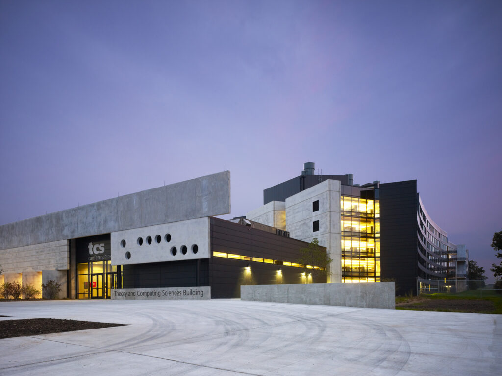 A modern concrete and glass building labeled "Thorvald Computing Sciences Building" with "tcs" logo, illuminated in the evening.