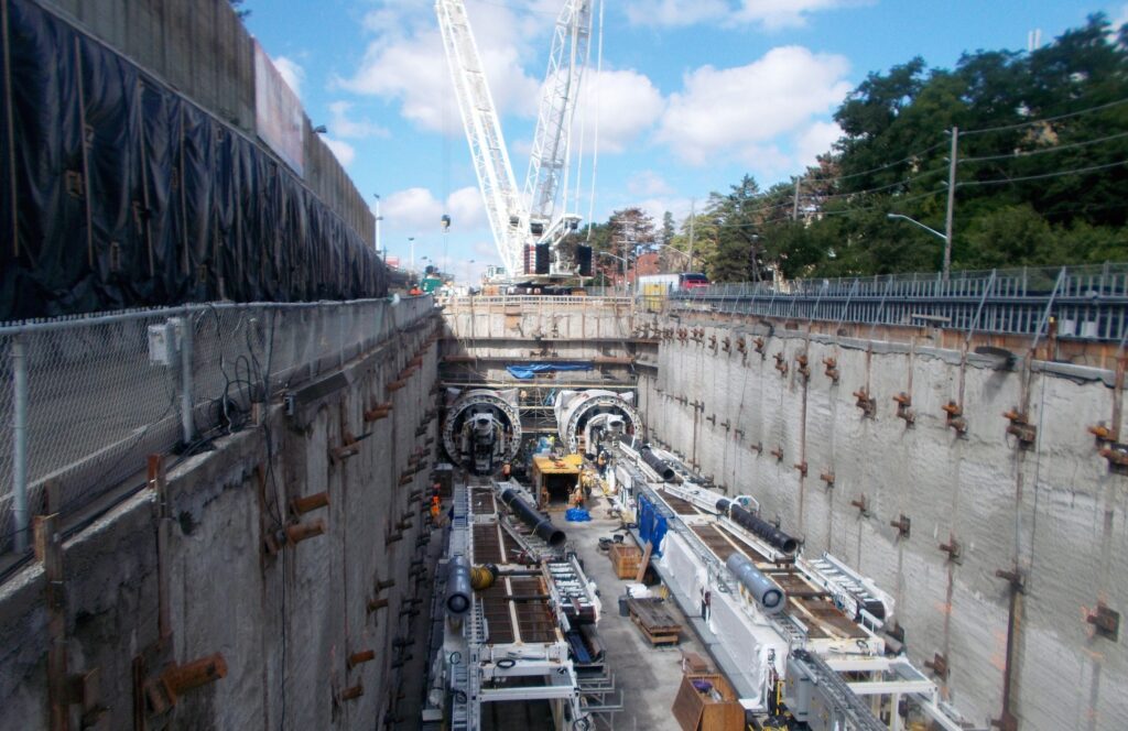 A large construction site with heavy machinery including cranes and tunnel boring machines, surrounded by reinforced concrete walls, and trees visible in the background.