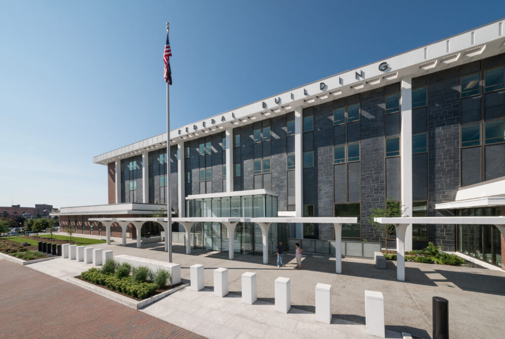 A modern office building with the word "FEDERAL BUILDING" above the entrance, an American flag on a pole in front, and a clear blue sky in the background.