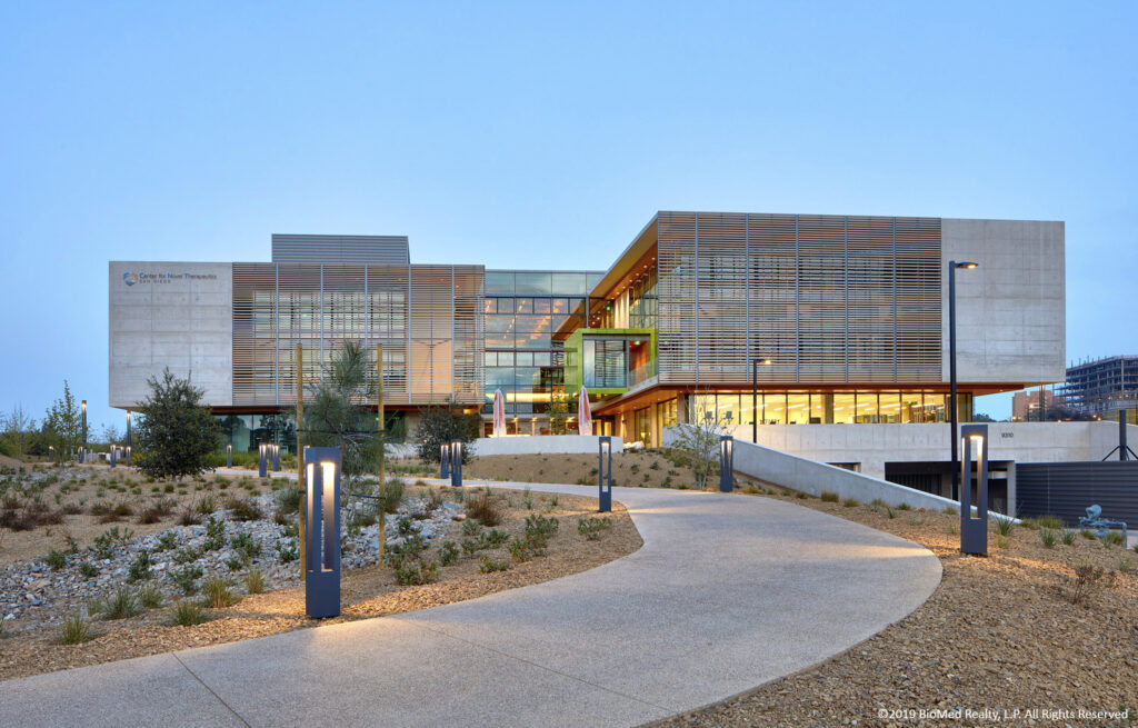 Modern building with large windows and horizontal slat exterior, located on a landscaped path with surrounding shrubbery and outdoor lighting.