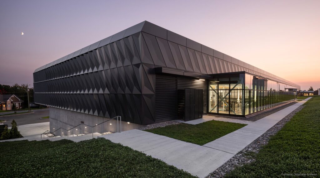 Modern architectural building with geometric black facade during twilight, featuring large glass windows and paths surrounding it, under a clear sky with a visible crescent moon.