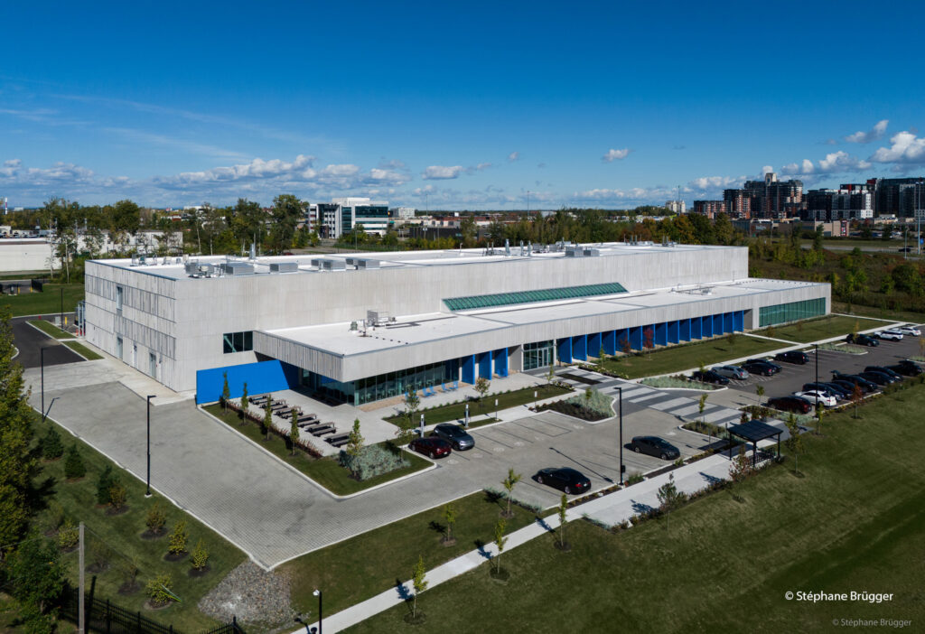 Aerial view of a large, modern white building with blue accents, surrounded by parking lots, green lawns, and trees under a clear sky.